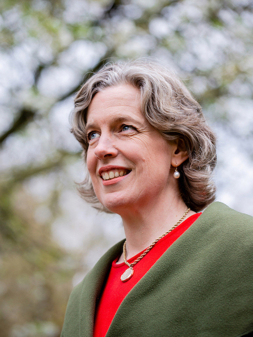 A shot taken from below of Emily Miles's head and shoulders. She wears a moss-green coat over a bright red dress and looks upwards at an angle. There are trees in the background